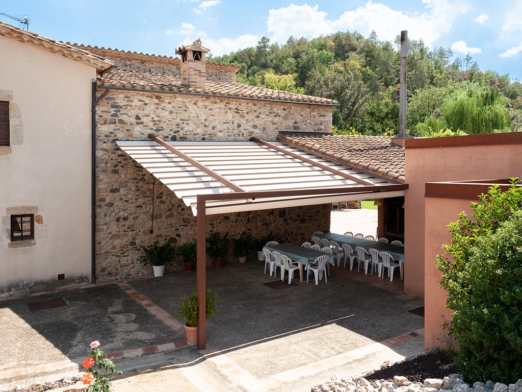 Taules i cadires sota una pèrgola, al fons hi ha una barbacoa argentina